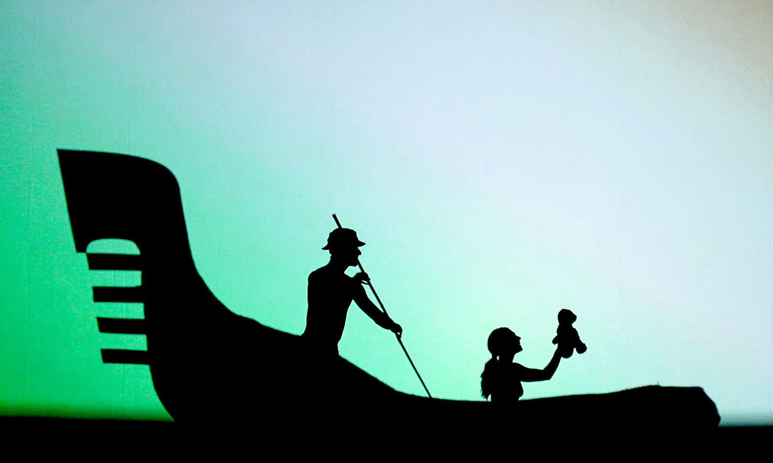 A shadow of a man rowing an Indigenous style canoe with a girl on board holding a teddy bear. 