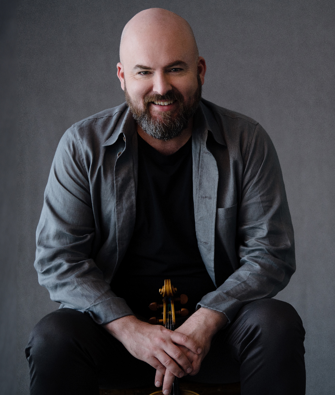 a bald white gentleman with a beard seated with his hands together on his lap, wearing a grey shirt