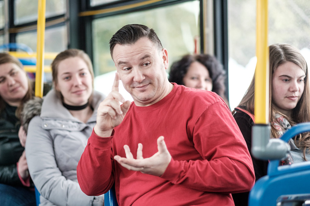 actor in a red shirt sits on a public transit bus