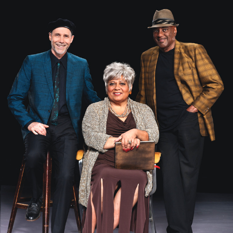 Two men standing and woman sitting on chair. 