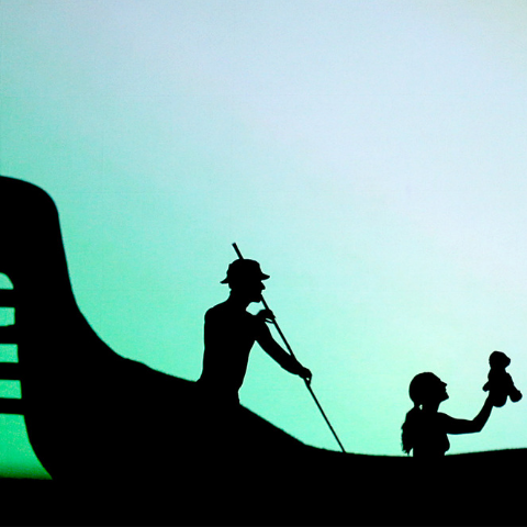 A shadow of a man rowing an Indigenous style canoe with a girl on board holding a teddy bear. 