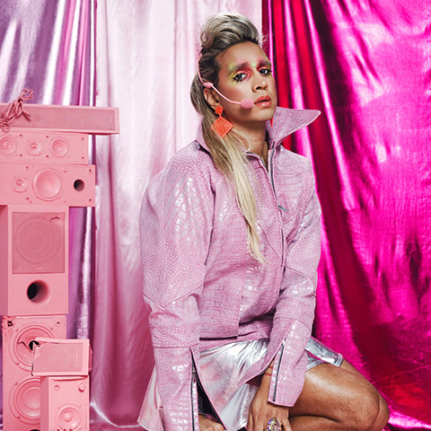 All pink, performer sits in a rain coat with wireless mic, furlong look on their face