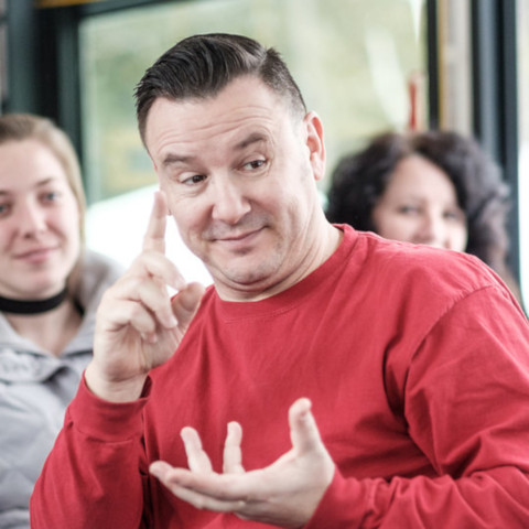 actor in a red shirt sits on a public transit bus